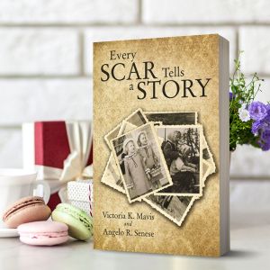 Paperback copy of Every Scar Tells a Story by Victoria K. Mavis and Angelo R. Senese stands upright. The sepia cover shows overlapping vintage photos of two little girls. Pastel macarons, a white coffee cup, and wrapped gifts sit beside it.