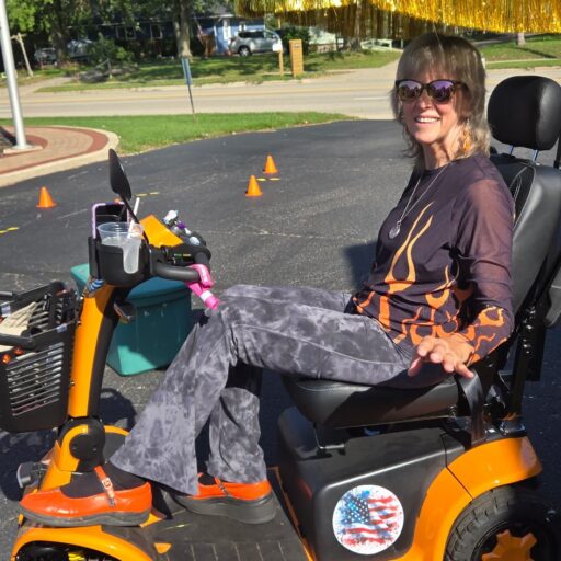 Victoria Mavis on her orange Harley-flame mobility scooter at the One Day in the Chair™ race course, surrounded by orange traffic cones in a parking lot. She is smiling and wearing a black and orange flame shirt — the same spirit as the race she created.